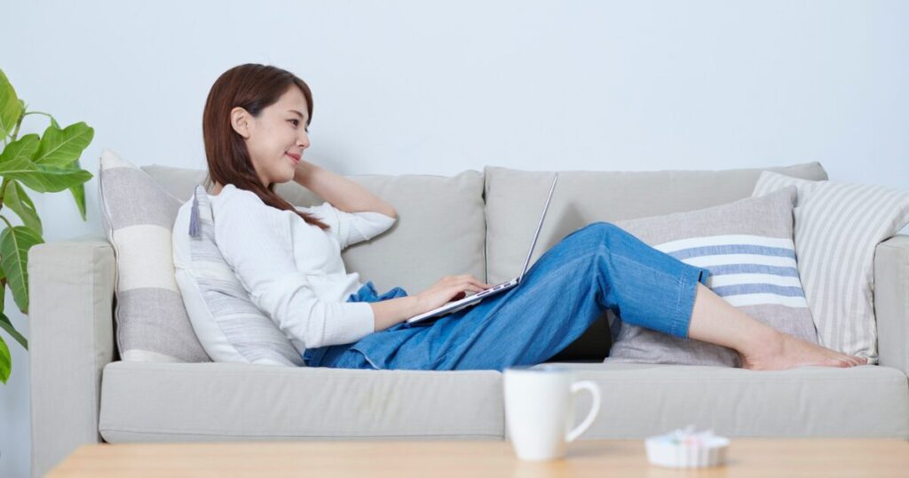 明るい部屋のソファでのんびりノートパソコンを見る女性
